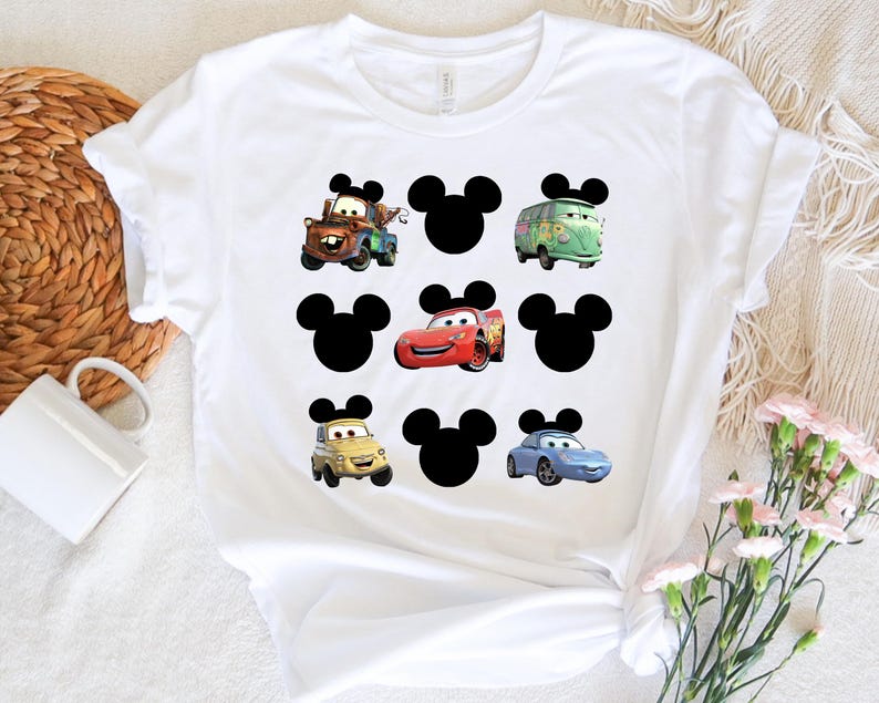 Pu&ograve; includere: Maglietta bianca con una grafica in bianco e nero di orecchie di Topolino e personaggi di Cars. I personaggi di Cars includono Mater, Saetta McQueen e un'auto da corsa blu.