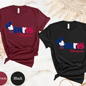 Puede incluir: Dos camisetas con un gr&aacute;fico de Mickey Mouse y el texto "Paris Disneyland". La camiseta de la izquierda es granate y la camiseta de la derecha es negra.