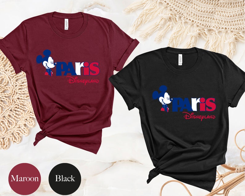 Puede incluir: Dos camisetas con un gr&aacute;fico de Mickey Mouse y el texto "Paris Disneyland". La camiseta de la izquierda es granate y la camiseta de la derecha es negra.