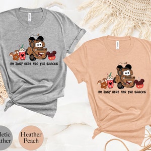 May include: Two t-shirts with a cartoon image of Mater from Cars, a Mickey Mouse ear hat, and snacks. The text on the shirts reads "I'm just here for the snacks". The shirts are available in athletic heather and heather peach.