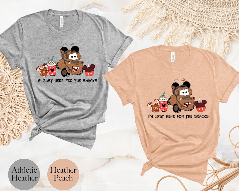 May include: Two t-shirts with a cartoon image of Mater from Cars, a Mickey Mouse ear hat, and snacks. The text on the shirts reads "I'm just here for the snacks". The shirts are available in athletic heather and heather peach.