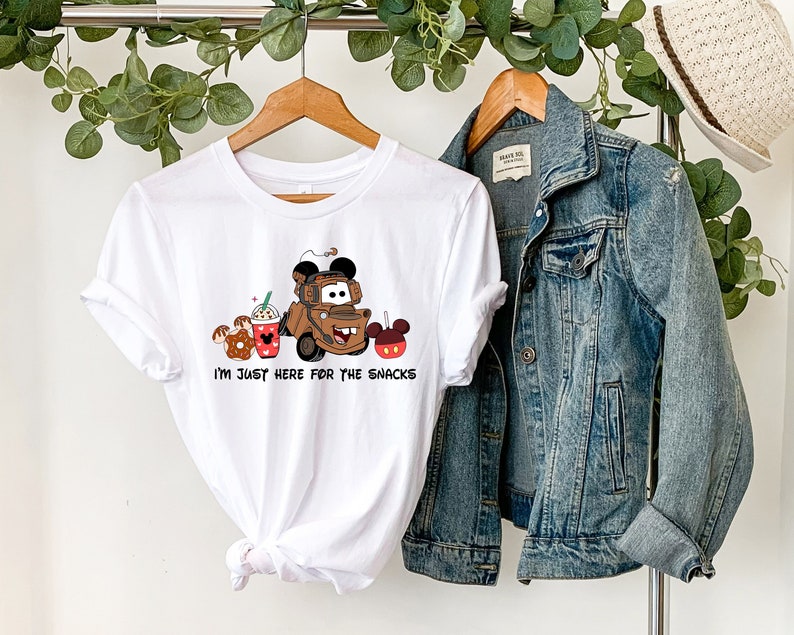 May include: White t-shirt with a brown cartoon truck and Mickey Mouse ears, surrounded by cartoon snacks. The text "I'm just here for the snacks" is printed below the image.