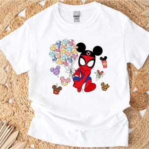 Puede incluir: Camiseta blanca con un personaje de dibujos animados de Spider-Man con orejas de Mickey Mouse. El personaje sostiene globos con forma de Mickey Mouse. Gr&aacute;ficos adicionales incluyen golosinas tem&aacute;ticas de Mickey Mouse en varios colores. La camiseta est&aacute; sobre un fondo tejido.