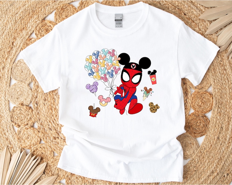 Puede incluir: Camiseta blanca con un personaje de dibujos animados de Spider-Man con orejas de Mickey Mouse. El personaje sostiene globos con forma de Mickey Mouse. Gr&aacute;ficos adicionales incluyen golosinas tem&aacute;ticas de Mickey Mouse en varios colores. La camiseta est&aacute; sobre un fondo tejido.