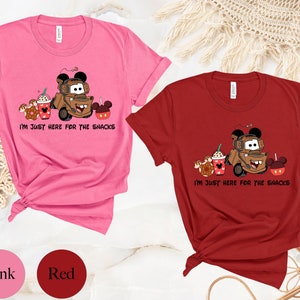 May include: Two t-shirts, one pink and one red, with a graphic of a brown tow truck with Mickey Mouse ears, holding a cup of coffee, a donut, and a Mickey Mouse pretzel. The text on the shirts reads "I'm just here for the snacks".