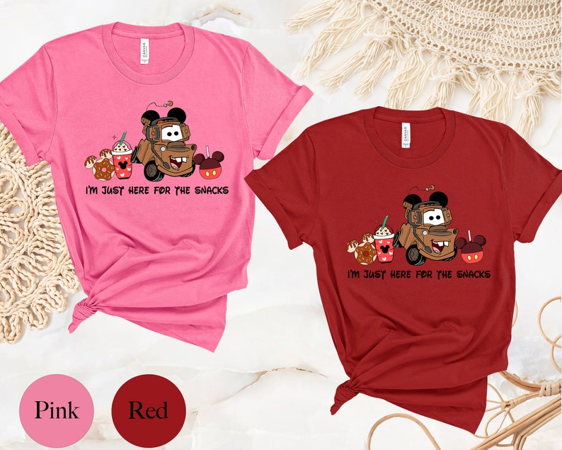 May include: Two t-shirts, one pink and one red, with a graphic of a brown tow truck with Mickey Mouse ears, holding a cup of coffee, a donut, and a Mickey Mouse pretzel. The text on the shirts reads "I'm just here for the snacks".