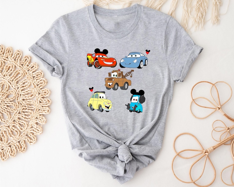 May include: Gray t-shirt with a graphic of five cartoon cars from the Disney Pixar movie Cars. The cars are red, blue, yellow, and brown. Each car has a Mickey Mouse ear on top.