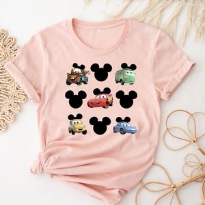 Pu&ograve; includere: Maglietta rosa con una silhouette nera di Topolino e immagini di personaggi di Cars, tra cui Saetta McQueen, Cricchetto e un'auto blu.