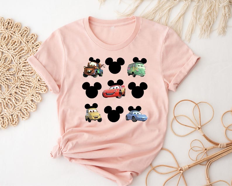 Pu&ograve; includere: Maglietta rosa con una silhouette nera di Topolino e immagini di personaggi di Cars, tra cui Saetta McQueen, Cricchetto e un'auto blu.