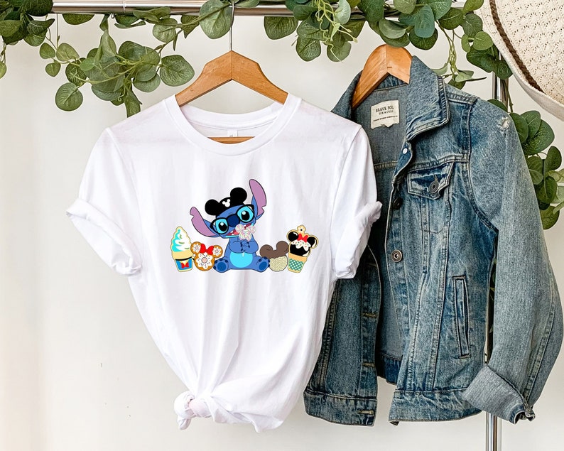 Pode incluir: Camiseta branca com um personagem de desenho animado usando &oacute;culos e gravata borboleta, cercado por sobremesas coloridas. O personagem &eacute; azul com orelhas grandes. Uma jaqueta jeans est&aacute; pendurada em um cabide ao lado da camiseta.