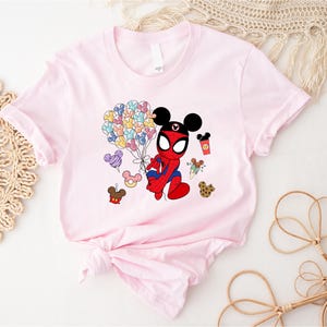 Puede incluir: Una camiseta rosa claro con un personaje de dibujos animados que combina a Spider-Man y Mickey Mouse. El personaje sostiene globos de colores y golosinas. La camiseta est&aacute; anudada en la parte inferior.