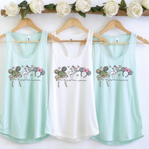 Pode incluir: Três blusas sem mangas em verde menta e branco penduradas em cabides de madeira. Cada top apresenta um gráfico de orelhas do Mickey Mouse, Olaf e um laço rosa, com o texto "For the first time in forever."