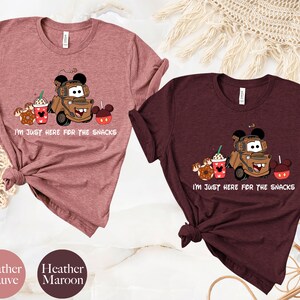 May include: Two t-shirts with a cartoon image of Mater from Cars, a donut, a coffee cup, and Mickey Mouse ears. The text on the shirts reads "I'm just here for the snacks". The shirts are heather mauve and heather maroon.