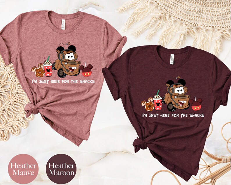May include: Two t-shirts with a cartoon image of Mater from Cars, a donut, a coffee cup, and Mickey Mouse ears. The text on the shirts reads "I'm just here for the snacks". The shirts are heather mauve and heather maroon.