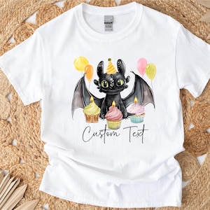 May include: White t-shirt featuring a black dragon character with green eyes, wearing a party hat, and surrounded by cupcakes and balloons. The text "Custom Text" is written below the dragon in a script font.