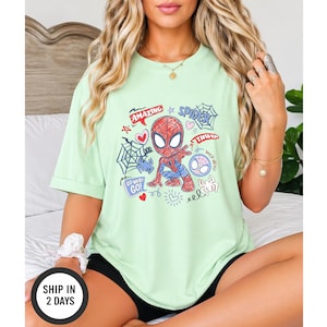 Puede incluir: Una camiseta verde claro con un dise&ntilde;o de dibujos animados de Spider-Man. El gr&aacute;fico incluye las palabras "Amazing", "Spider" y "Go!" junto con corazones, una telara&ntilde;a y otros elementos decorativos. La camiseta es de estilo casual.