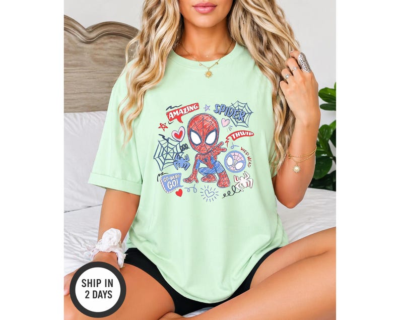 Puede incluir: Una camiseta verde claro con un dise&ntilde;o de dibujos animados de Spider-Man. El gr&aacute;fico incluye las palabras "Amazing", "Spider" y "Go!" junto con corazones, una telara&ntilde;a y otros elementos decorativos. La camiseta es de estilo casual.