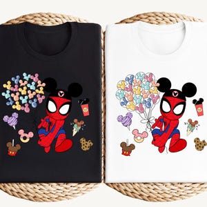 Puede incluir: Camisetas negras y blancas con un personaje de dibujos animados de Spider-Man con orejas de Mickey Mouse. El dise&ntilde;o incluye globos de colores con forma de Mickey Mouse, helados, donuts y tazas de caf&eacute;. Las camisetas se muestran sobre una superficie tejida.