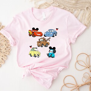 May include: Pink t-shirt with a graphic of five cartoon cars from the Disney Pixar movie Cars. The cars include Lightning McQueen, Mater, and a blue car with a Mickey Mouse ear hat.
