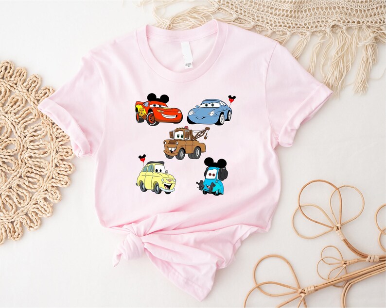May include: Pink t-shirt with a graphic of five cartoon cars from the Disney Pixar movie Cars. The cars include Lightning McQueen, Mater, and a blue car with a Mickey Mouse ear hat.