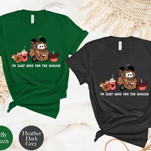 May include: Two t-shirts with a cartoon image of Mater from Cars, a Mickey Mouse head, and snacks. The text on the shirts reads "I'm just here for the snacks". The shirts are available in Kelly Green and Heather Dark Grey.