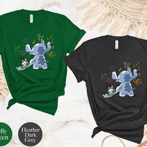 May include: Two t-shirts featuring a blue Stitch character from Lilo & Stitch. The Stitch character is holding a pencil and drawing on a wall. The t-shirts are available in Kelly Green and Heather Dark Grey.