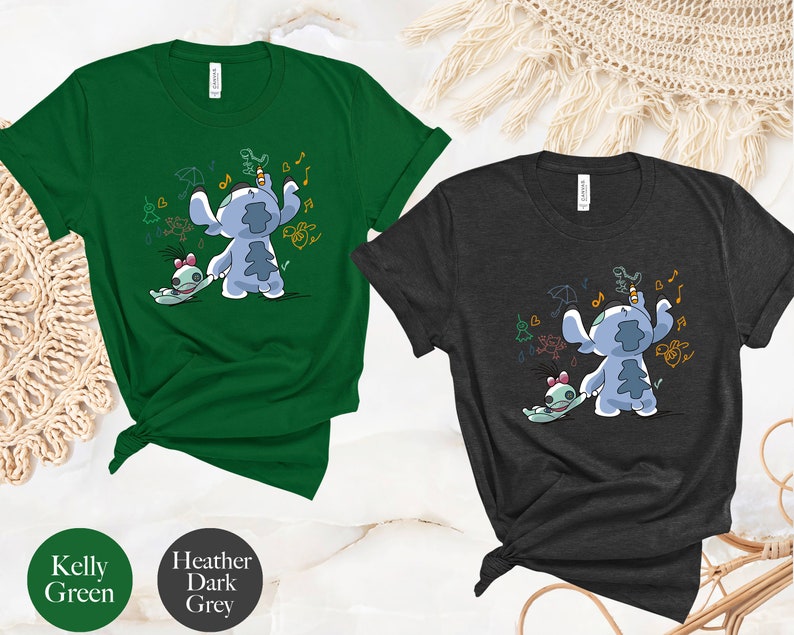 May include: Two t-shirts featuring a blue Stitch character from Lilo & Stitch. The Stitch character is holding a pencil and drawing on a wall. The t-shirts are available in Kelly Green and Heather Dark Grey.