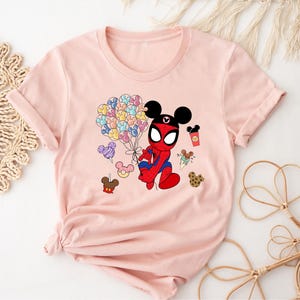 Puede incluir: Camiseta rosa claro con un personaje de dibujos animados que combina a Spider-Man y Mickey Mouse. El personaje sostiene globos y una taza de caf&eacute;. Otras im&aacute;genes de dibujos animados tambi&eacute;n est&aacute;n impresas en la camiseta.