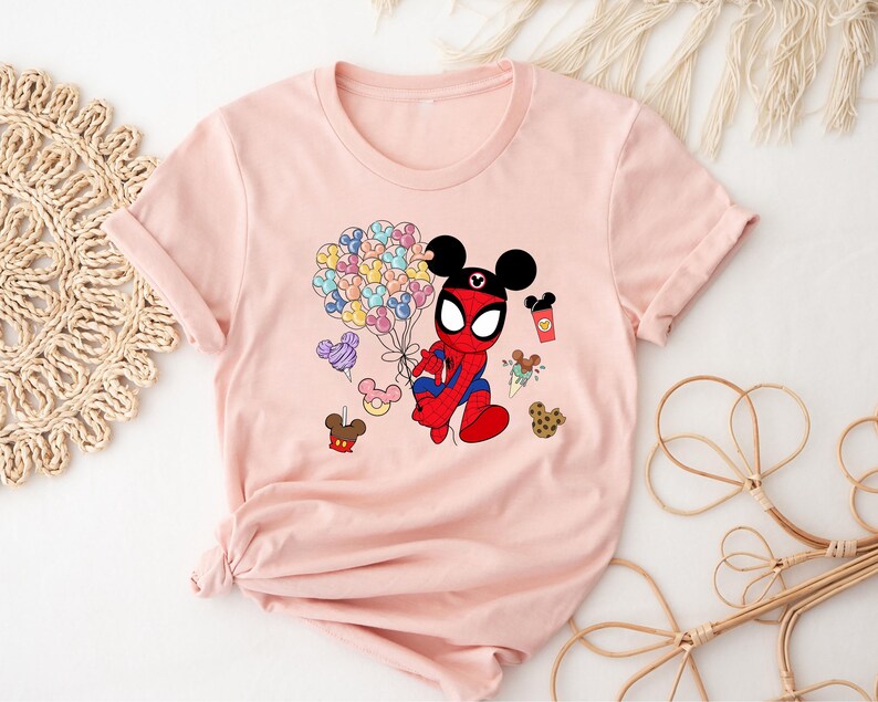 Puede incluir: Camiseta rosa claro con un personaje de dibujos animados que combina a Spider-Man y Mickey Mouse. El personaje sostiene globos y una taza de caf&eacute;. Otras im&aacute;genes de dibujos animados tambi&eacute;n est&aacute;n impresas en la camiseta.