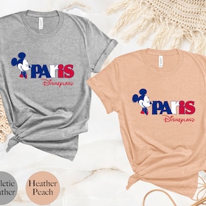 Puede incluir: Dos camisetas con un gr&aacute;fico de Mickey Mouse y el texto "Paris Disneyland". La camiseta de la izquierda es gris deportivo jaspeado y la camiseta de la derecha es melocot&oacute;n jaspeado.