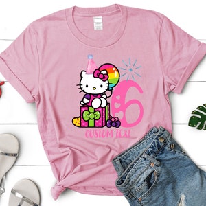 May include: Pink t-shirt with a cartoon image of Hello Kitty wearing a party hat and sitting on a gift box with a balloon and the number 6. The text "CUSTOM TEXT" is below the image.