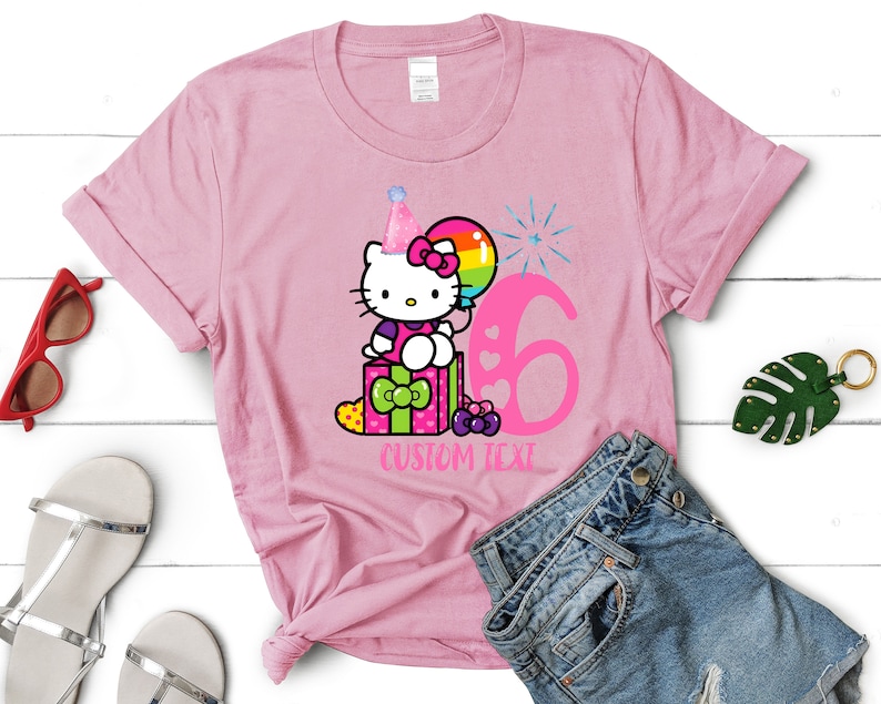 May include: Pink t-shirt with a cartoon image of Hello Kitty wearing a party hat and sitting on a gift box with a balloon and the number 6. The text "CUSTOM TEXT" is below the image.