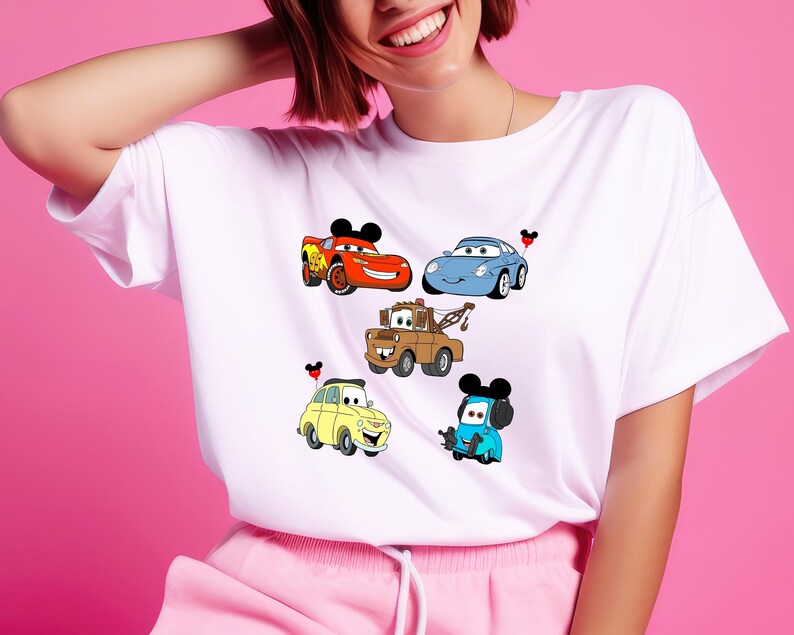 May include: White t-shirt with a colorful cartoon design of five cars from the Disney Pixar movie Cars. The cars are red, blue, yellow, brown, and turquoise. Each car has a Mickey Mouse ear on top.