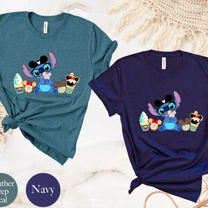 Pode incluir: Duas camisetas, uma verde-azulado escuro e outra azul marinho, cada uma com um personagem de desenho animado Stitch usando &oacute;culos e um chap&eacute;u com orelhas do Mickey Mouse. O personagem &eacute; cercado por sobremesas, incluindo sorvetes e cupcakes.