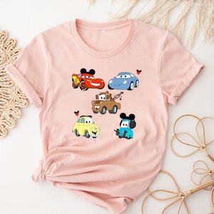 May include: Pink t-shirt with a graphic of five cartoon cars from the Disney Pixar movie Cars. The cars are Lightning McQueen, Mater,  a blue car, a yellow car, and a red car. Each car has a Mickey Mouse ear on top.