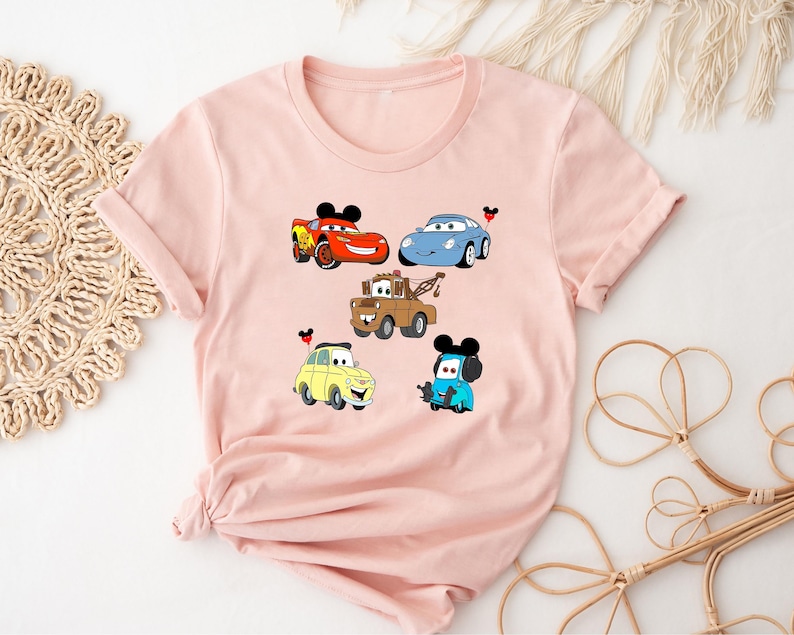 May include: Pink t-shirt with a graphic of five cartoon cars from the Disney Pixar movie Cars. The cars are Lightning McQueen, Mater,  a blue car, a yellow car, and a red car. Each car has a Mickey Mouse ear on top.