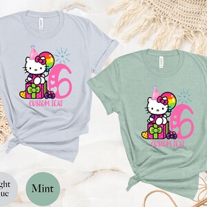 May include: Two t-shirts with a Hello Kitty design. The light blue shirt has a pink number 6 and the mint green shirt has a pink number 6. Both shirts have a Hello Kitty character holding a balloon and sitting on a gift box. The text "CUSTOM TEXT" is below the design on both shirts.