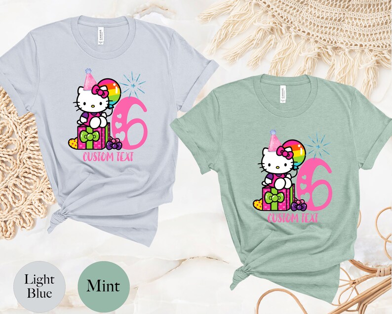 May include: Two t-shirts with a Hello Kitty design. The light blue shirt has a pink number 6 and the mint green shirt has a pink number 6. Both shirts have a Hello Kitty character holding a balloon and sitting on a gift box. The text "CUSTOM TEXT" is below the design on both shirts.