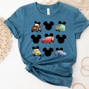Pu&ograve; includere: Una maglietta blu turchese con una stampa grafica di orecchie di Topolino e personaggi di Cars. I personaggi di Cars includono Saetta McQueen, Cricchetto e un'auto da corsa blu.