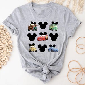 Pu&ograve; includere: Maglietta grigia con un design di Topolino con vari personaggi di Cars, tra cui Saetta McQueen, Cricchetto e Cricchetto il carro attrezzi.