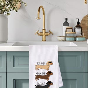 May include: White waffle-weave kitchen towel featuring three dachshund illustrations, each wearing sunglasses. The towel has the text "LIGHT ROAST", "MEDIUM ROAST", and "DARK ROAST" above each dog. The towel is hanging on a blue cabinet.