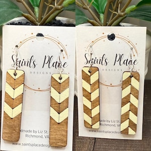 May include: Two pairs of wooden earrings with a chevron design. The earrings are rectangular with a light brown base and a cream-colored chevron pattern. The earrings are displayed on cards with the text "Saints Place Designs."