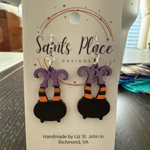 May include: A pair of Halloween-themed earrings featuring a black cauldron design with purple witch legs and orange and black striped socks. The earrings are handmade and have silver-colored hooks. The card behind the earrings says "Saints Place Designs."