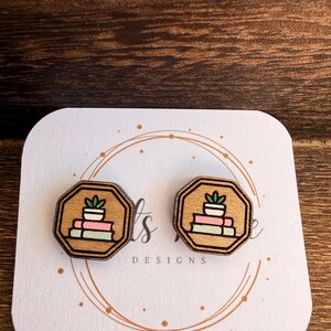 May include: Wooden stud earrings featuring a stack of books with a potted plant on top. The earrings have a light brown background with a dark brown octagonal border. The earrings are on a white card with the text "ts DESIGNS".