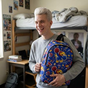 May include: A blue backpack featuring a repeating pattern of a cartoon bird mascot holding red pom-poms, interspersed with yellow stars, red megaphones, and swirling white lines. The backpack has a rounded top, a front pocket, and black straps.