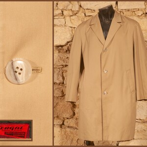 Puede incluir: Una gabardina de color tostado con cuello de solapa, mangas largas y cierre frontal de tres botones. La imagen incluye un primer plano de un botón redondo de color claro y una etiqueta de tela roja con el texto Tergal Marque Deposee Fibre Polyester Melange.