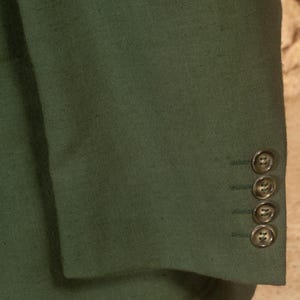 May include: Close-up of a dark green blazer sleeve. The fabric has a textured appearance. Four matching buttons are visible on the cuff. The blazer is a solid color, and the buttons are a similar shade of green.