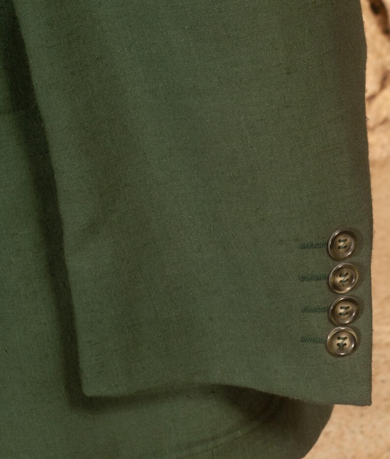 May include: Close-up of a dark green blazer sleeve. The fabric has a textured appearance. Four matching buttons are visible on the cuff. The blazer is a solid color, and the buttons are a similar shade of green.