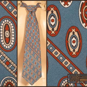 May include: A blue necktie featuring a repeating pattern of red, white, and dark blue oval motifs. The design includes diagonal bands with geometric square and circle shapes. A small rectangular fabric label is attached to the lower right corner.