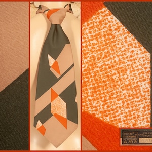 Puede incluir: Una corbata con un patrón geométrico en verde oliva oscuro, beige y naranja brillante. El diseño incluye bloques sólidos y formas de diamante texturizadas. Una pequeña etiqueta rectangular en la parte inferior dice "TERITAL" y "100% POLYESTER".